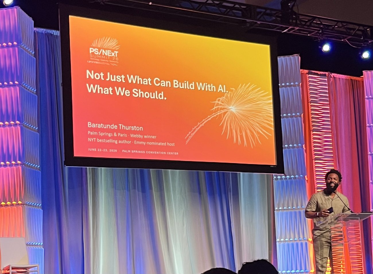 Baratunde Thurston speaks at the PS/NExT Summit 2026 beneath a slide reading Not Just What We Can Build With AI. What We Should.