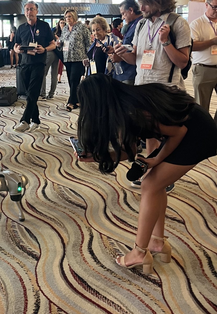 KMIR/NBC Palm Springs anchor Alondra Campos films a robot dog with her station microphone in the Palm Springs Convention Center lobby at PS/NExT Summit 2026.
