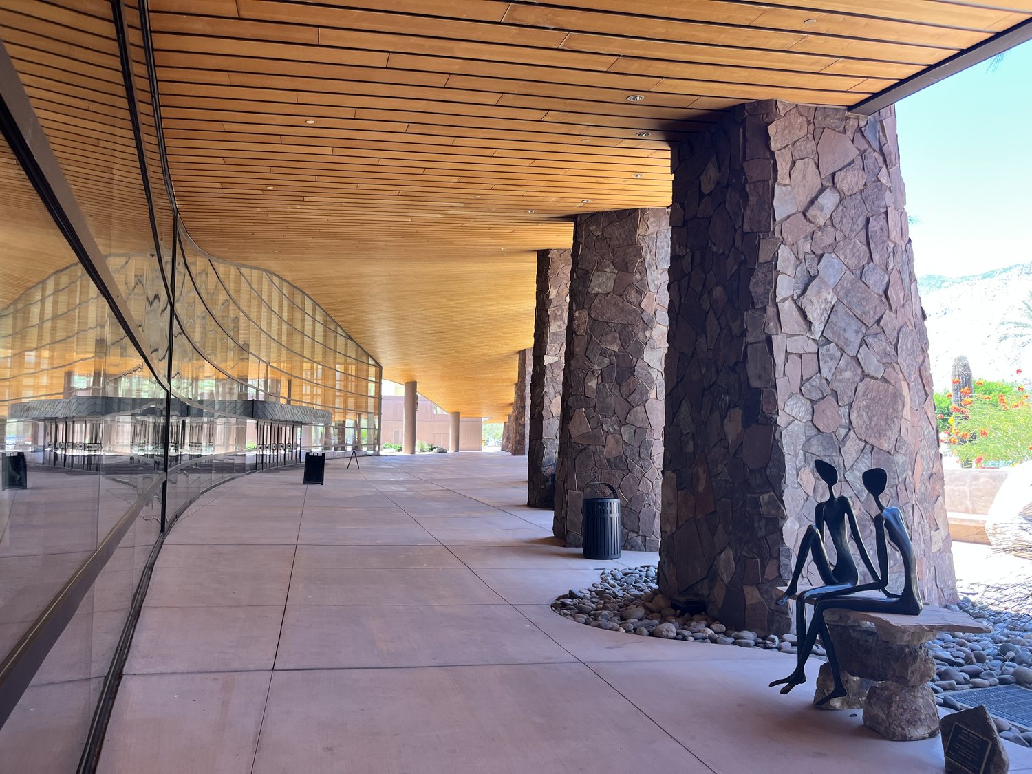 The wood-and-stone colonnade of the Palm Springs Convention Center, two bronze figures seated together beneath a desert-stone pillar — site of the PS/NExT Summit 2026.